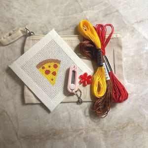 May include: A needlepoint kit featuring a canvas with a pizza slice design, a beige zippered pouch, three skeins of embroidery floss in yellow, red, and brown, a small pink thread cutter, a red flower-shaped needle minder, and a silver needle.