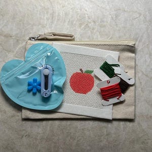 May include: A beige canvas zipper pouch containing a white needlepoint canvas with a red apple design, three cardboard bobbins wrapped in red, green, and brown embroidery thread, and a light blue heart-shaped pouch holding a blue flower needle threader.