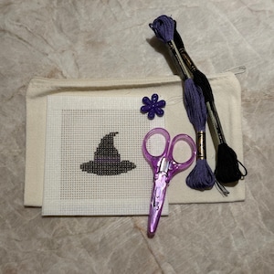 May include: A cross-stitch kit featuring a square white Aida cloth with a stitched black witch hat, a beige canvas zipper pouch, purple and black embroidery floss, a pair of translucent purple scissors, and a small purple flower-shaped button.