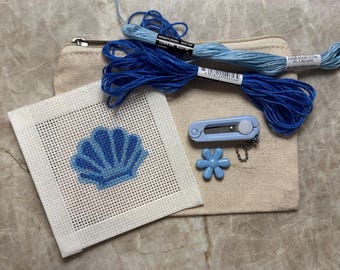 May include: A needlepoint canvas featuring a blue seashell design, two skeins of blue embroidery floss, a light blue thread cutter, and a small light blue flower charm, all resting on a beige canvas zipper pouch.