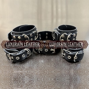 May include: Black leather restraints with silver metal hardware. The set includes wrist cuffs, an ankle cuff, and a neck collar. Each piece features rivets and adjustable buckles. The brand name "LUXGRAIN LEATHER" is printed on the image.