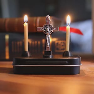 May include: A small, black metal box holds a silver crucifix and two lit candles. The crucifix depicts Jesus on the cross. The candles are lit with yellow flames. The background includes a stack of books.