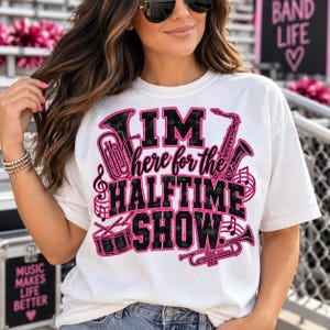May include: A white short-sleeve t-shirt featuring a graphic design with the text "I'M here for the HALFTIME SHOW." The design includes black and pink illustrations of a tuba, saxophone, drum, and trumpet, along with musical notes.