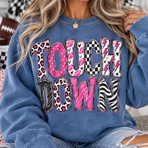 Puede incluir: Una sudadera azul de cuello redondo con el texto TOUCH DOWN en letras grandes con varios patrones. Las letras incluyen balones de fútbol americano rosas y blancos, estampado de leopardo, cuadros, rayos rosas y rayas de cebra en blanco y negro.