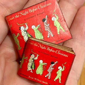 May include: Two miniature red books with the title "Twas the Night Before Christmas" and illustrations of figures in pajamas. The books are held in a hand, showcasing their small size and vintage appearance. The author's name, Clement Moore, is also visible.