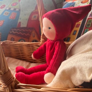 Puede incluir: Una muñeca de fieltro rojo con una capucha puntiaguda se sienta en una cesta de mimbre. La muñeca tiene una cara simple con ojos negros y una sonrisa cosida. La muñeca lleva un atuendo de fieltro rojo.