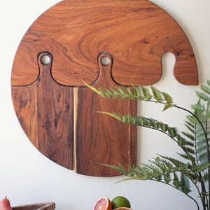 May include: A set of wooden cutting boards in various shapes and sizes. The largest board is circular, with three smaller boards attached. A small rectangular board with a handle rests on a woven basket, with sliced citrus fruit.