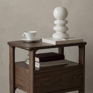 May include: A dark brown wooden bedside table with a lower drawer and a shelf. A white teacup and saucer sit on top, along with books and a white sculpture. The table has rounded edges and legs.