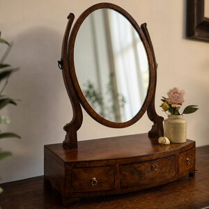 Op de afbeelding: Een houten toilettafelspiegel met een ovale spiegel en een gebogen voet met drie lades. De spiegel wordt ondersteund door sierlijke, gebogen houten armen. Een kleine vaas met bloemen en een zeeschelp staan op de toilettafel. Het hout heeft een rijke, donkerbruine afwerking.