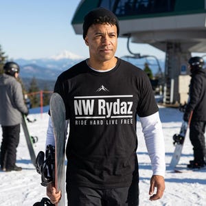 May include: A black t-shirt with the text "NW Rydaz" and "Ride Hard Live Free" in white. The shirt is worn over a white long-sleeve shirt. A person is holding a snowboard on a snowy mountain with other snowboarders in the background.