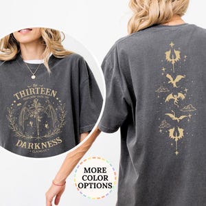 May include: A charcoal gray short-sleeve t-shirt featuring a gold graphic on the back with a vertical arrangement of dragon silhouettes and stars. The front chest area displays a circular gold design with wings, a sword, and the text THE THIRTEEN.