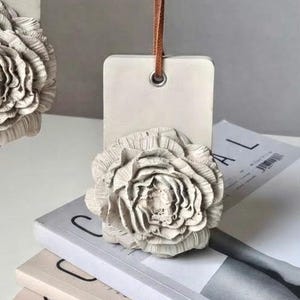 Pode incluir: Uma etiqueta retangular de gesso em tom branco sujo com uma flor esculpida em três dimensões na parte frontal. A etiqueta possui um ilhó metálico no topo e está suspensa por um fino cordão de couro castanho. As pétalas são detalhadas.