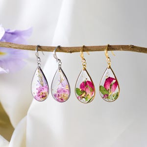 May include: Two pairs of teardrop-shaped resin earrings hanging from a thin branch. One pair features silver-toned hooks and frames with purple pressed flowers and gold flakes. The other pair has gold-toned hooks and frames with pink flowers and green leaves.
