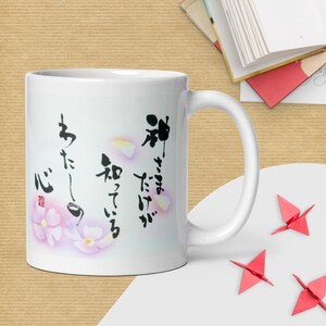 以下が含まれることがあります： 黒い日本語の書道文字と淡いピンクの花のイラストが描かれた白いセラミック製のマグカップ。左下には小さな赤い四角い印があり、筆文字の周りには繊細なピンクの花びらが散りばめられたデザインになっています。
