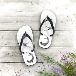 May include: White flip-flops with black straps, each featuring a detailed black and white illustration of a fish. The sandals are arranged on a weathered, light-colored wooden surface, suggesting a beach or outdoor theme.