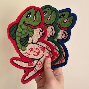 May include: Three embroidered fish-shaped coasters in green, red, and blue. The fish have green scales, red lips, and tan bellies with red anchor and floral patterns. The coasters are held in a hand.