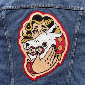 May include: A denim jacket with a large, colourful patch. The patch features a woman's face with gold hair, a white wolf head, and a hand. The patch has a red border with gold and red accents.