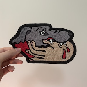 May include: Embroidered patch featuring a gray dog biting a tan hand with red blood drops. The patch has a black border and is likely for clothing or accessories.