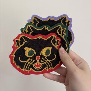 May include: A stack of three embroidered cat face patches. Each patch features a black cat face with gold thread outlines, green eyes, and a red nose and mouth. The patches have distinct red, dark green, and light purple felt backing borders.