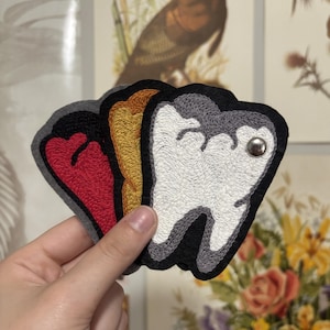 May include: A set of three embroidered tooth-shaped patches featuring a black border and a metallic snap button on the white patch. The patches are layered, displaying a white, a mustard yellow, and a red tooth design, each with grey stitching.