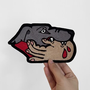 Puede incluir: Un parche bordado que muestra una cabeza de perro gris con un contorno negro, posicionado sobre una mano de color beige. La mano tiene uñas rosas y dos gotas rojas que parecen sangre. El parche tiene un borde negro grueso y un área roja.