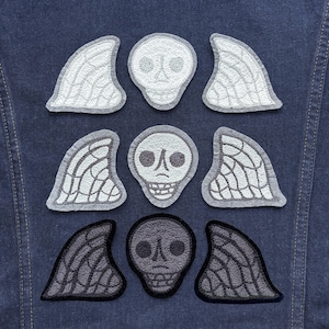 May include: Three rows of embroidered patches arranged on dark blue denim fabric. Each row features a central skull flanked by two wing-like shapes with intricate webbed patterns. The top two rows are light gray, while the bottom row is black.