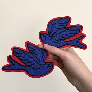 May include: Two blue bird patches with red borders. The birds are embroidered with black details. These patches can be used to decorate clothing, bags, or other fabric items. The patches are held up against a white background.