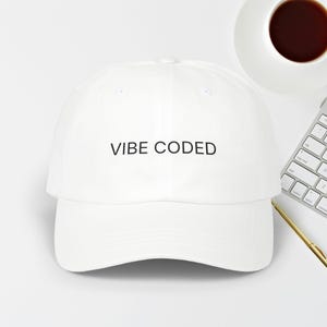 May include: A white baseball cap with the words "VIBE CODED" printed in black. The cap is set against a white background, with a coffee cup and keyboard visible.