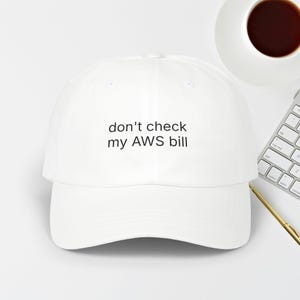 May include: A white baseball cap with the text "don't check my AWS bill" in black. The cap is on a white surface, with a cup of coffee and a keyboard in the background.