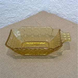 May include: A rectangular amber-colored glass dish featuring a pressed diamond and starburst pattern across its exterior walls and a small handle on one side. The base of the dish displays a radiating sunburst design etched into the glass.