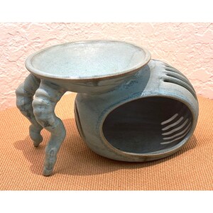 May include: A light blue ceramic oil burner with a smooth, rounded body and a wide, shallow basin on top. The base features a large oval opening and is supported by two curved, textured legs that extend outward from the side of the structure.