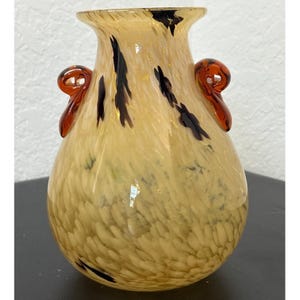 May include: A glass vase with a bulbous body and a flared rim, featuring a speckled cream and tan pattern with dark brown streaks. The vase includes two decorative, curled amber-colored glass handles attached to the upper sides.