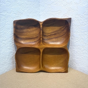 May include: A square wooden serving tray featuring four distinct, rounded compartments. The tray is crafted from a medium-brown wood with a visible, swirling grain pattern throughout the surface, showcasing natural variations in color and texture.