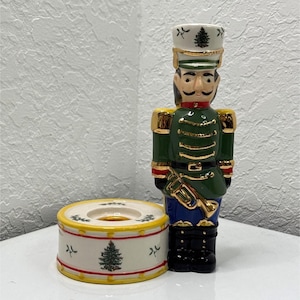May include: A ceramic nutcracker figurine standing next to a cylindrical drum-shaped candle holder. The nutcracker wears a green uniform with gold trim, a white hat with a tree design, and black boots. The white drum features red stripes and trees.