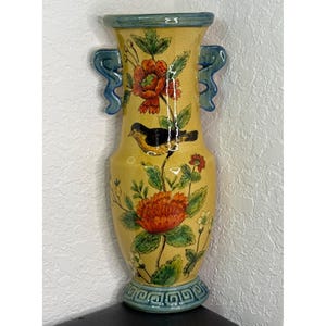 May include: A tall yellow ceramic vase featuring two blue handles and a hand-painted design of a black bird perched on a branch surrounded by large orange and red flowers with green leaves. The base and rim are decorated with a blue geometric pattern.