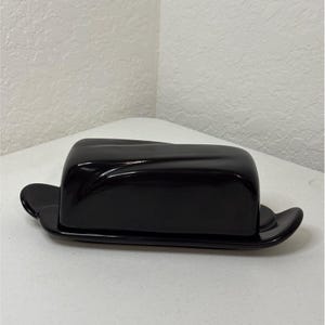 May include: A solid black butter dish featuring a rectangular base with extended handles on each side and a matching rectangular lid with a smooth, glossy finish and subtle, molded diagonal indentations across the top surface.