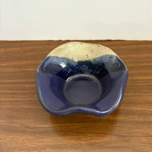 May include: A small, handmade ceramic dish with a unique shape. The dish features a deep blue interior and a light beige rim. The exterior is a glossy finish, perfect for holding small items.