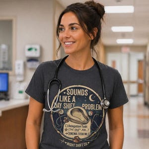 May include: A dark grey t-shirt with the text "Sounds Like a Day Shift Problem" and a medical graphic. The shirt also features the text "Night Shift Co." and a delegation task. The person is wearing blue scrubs and a stethoscope.
