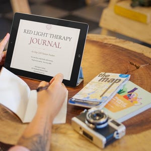 May include: A digital tablet displays the text "RED LIGHT THERAPY JOURNAL" with additional text. A notebook is open, and a pen is poised above the blank pages. A camera and two books are on a wooden table.