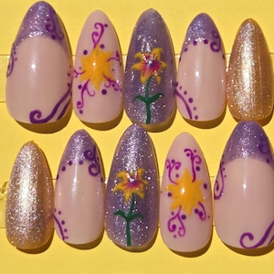 May include: A set of ten almond-shaped artificial nails. The nails feature a variety of designs, including glitter, floral patterns, and purple accents. Some nails have a nude base with purple swirls and dots. Others have a gold glitter finish.