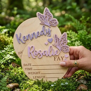 Puede incluir: Dos letreros de madera para anunciar el nacimiento de un bebé con los nombres "Kennedy" y "Rosalie" en escritura lavanda. Cada letrero tiene mariposas y corazones lavanda. El letrero inferior incluye líneas para fecha, hora, peso y longitud.