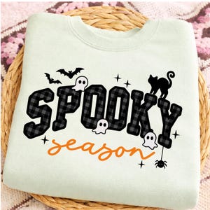 May include: A light green crewneck sweatshirt featuring the text SPOOKY in a black and gray buffalo plaid pattern and the word season in orange script. The design includes black silhouettes of two bats, a cat, a spider, and three white ghosts.