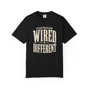 May include: Black t-shirt with the words "ELECTRICIAN WIRED DIFFERENT" in a distressed, off-white font. A lightning bolt graphic is between the words "WIRED" and "DIFFERENT". The shirt has short sleeves and a crew neck.