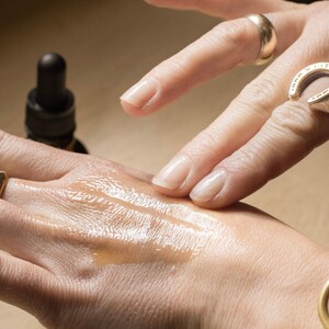 May include: A close-up of a hand with a liquid substance being applied. The liquid is a light amber color and appears to be a serum or oil. A dark brown bottle with a dropper is visible in the background. The hand is adorned with gold rings and a gold bracelet.