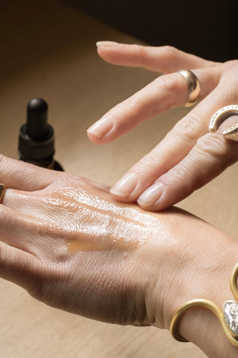 May include: A close-up of a hand with a liquid substance being applied. The liquid is a light amber color and appears to be a serum or oil. A dark brown bottle with a dropper is visible in the background. The hand is adorned with gold rings and a gold bracelet.