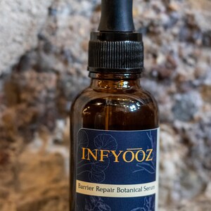 May include: A brown glass bottle of INFYOOZ Barrier Repair Botanical Serum with a black dropper. The label is navy blue with gold text and botanical illustrations. The bottle contains 1 oz of serum.
