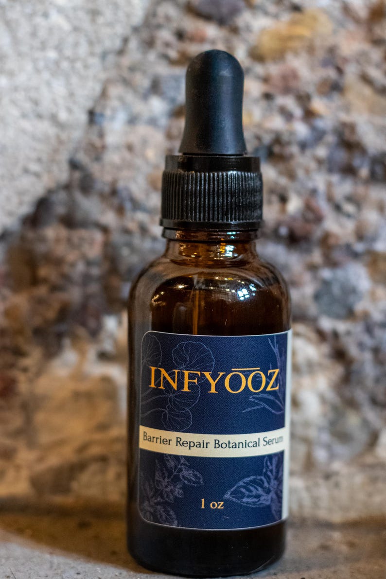 May include: A brown glass bottle of INFYOOZ Barrier Repair Botanical Serum with a black dropper. The label is navy blue with gold text and botanical illustrations. The bottle contains 1 oz of serum.