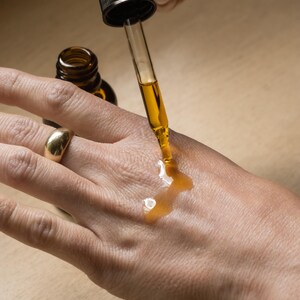 May include: A clear glass dropper dispenses a golden liquid onto a person's hand. The dropper is attached to a dark brown bottle. A gold ring adorns the person's finger. The image suggests skincare or cosmetic products.