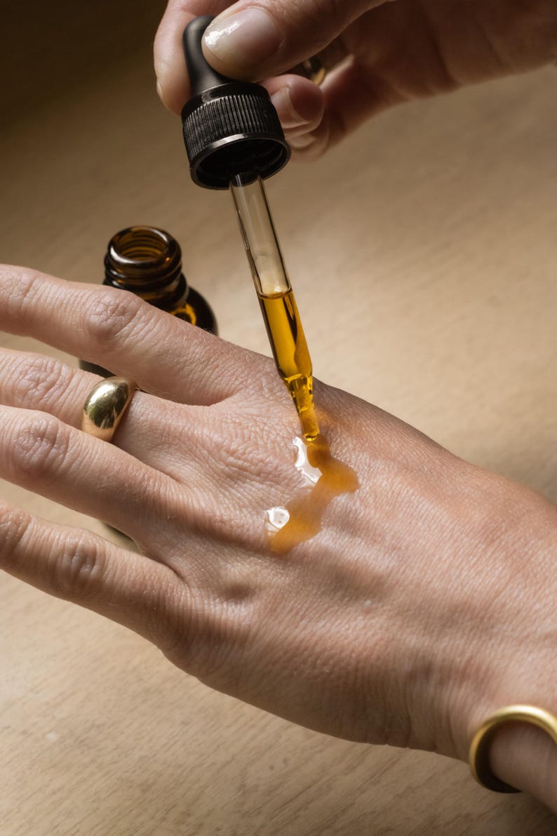 May include: A clear glass dropper dispenses a golden liquid onto a person's hand. The dropper is attached to a dark brown bottle. A gold ring adorns the person's finger. The image suggests skincare or cosmetic products.