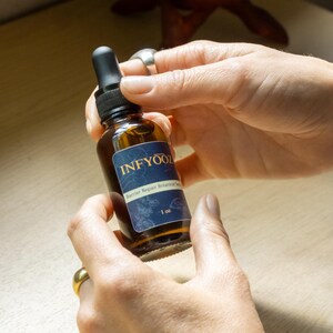 May include: A brown glass bottle with a black dropper is held between two hands. The bottle has a blue label with the text "INFYOOZ" and "Barrier Repair Botanical Serum". The label also indicates the bottle contains 1 oz of product.